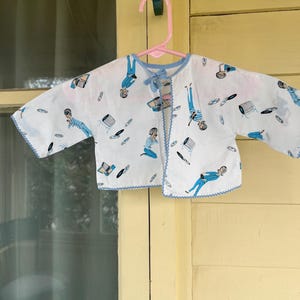 May include: A white baby jacket with a blue trim and a pattern of figures, records, and record players. The jacket is hanging on a pink hanger against a yellow background.