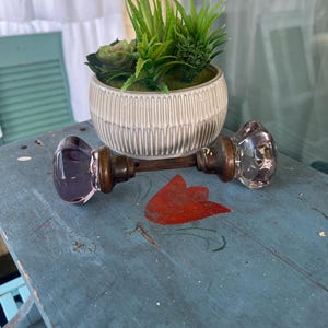 May include: A vintage door knob set featuring two faceted, light purple glass knobs connected by a central metal spindle with a dark bronze finish. The knobs have a clear, multi-faceted design that catches light, attached to circular metal bases.