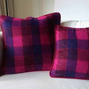 K&ouml;nnte beinhalten: Zwei dekorative Kissen mit einem pink- und lilafarbenen Karomuster. Die Kissen liegen auf einem wei&szlig;en Sofa.