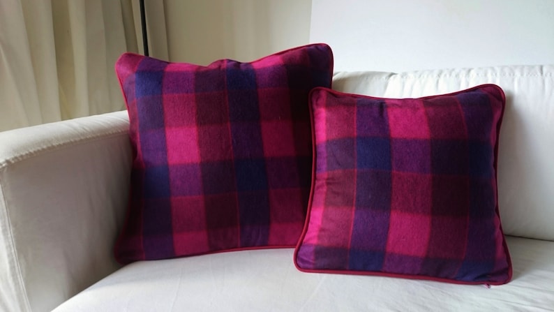 K&ouml;nnte beinhalten: Zwei dekorative Kissen mit einem pink- und lilafarbenen Karomuster. Die Kissen liegen auf einem wei&szlig;en Sofa.
