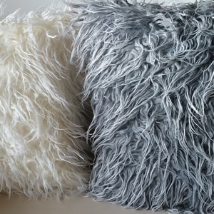 Puede incluir: Dos almohadas mullidas, una blanca y otra gris, est&aacute;n dispuestas en un sof&aacute;. Las almohadas est&aacute;n hechas de un material suave y peludo.