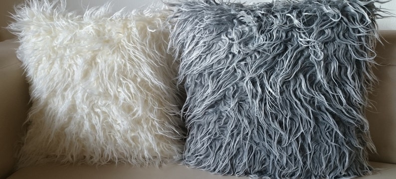 Puede incluir: Dos almohadas mullidas, una blanca y otra gris, est&aacute;n dispuestas en un sof&aacute;. Las almohadas est&aacute;n hechas de un material suave y peludo.