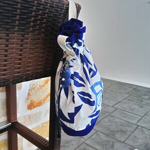 May include: A white and blue drawstring bag with a blue floral pattern. The bag is hanging on a brown wicker chair. A blue and white beaded charm hangs from the bag.