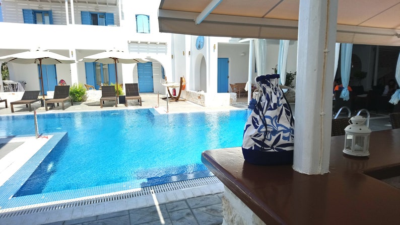 May include: A blue and white patterned drawstring bag sits on a wooden counter next to a white lantern. A swimming pool and white buildings are visible in the background.