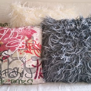 Puede incluir: Tres almohadas decorativas. Una almohada es blanca y esponjosa, otra es gris y esponjosa, y la tercera es una almohada rectangular con fondo blanco y dise&ntilde;os de estilo graffiti coloridos.