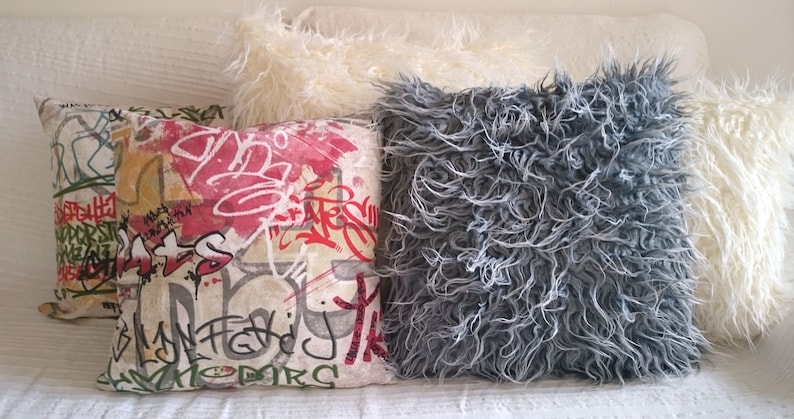 Puede incluir: Tres almohadas decorativas. Una almohada es blanca y esponjosa, otra es gris y esponjosa, y la tercera es una almohada rectangular con fondo blanco y dise&ntilde;os de estilo graffiti coloridos.