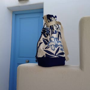 May include: A white canvas drawstring bag with blue floral print and a blue trim. The bag has a beige drawstring and a silver charm.