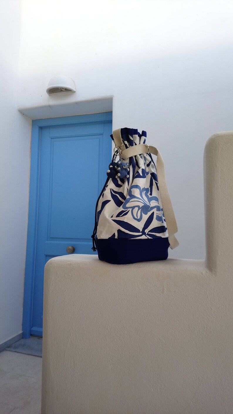 May include: A white canvas drawstring bag with blue floral print and a blue trim. The bag has a beige drawstring and a silver charm.