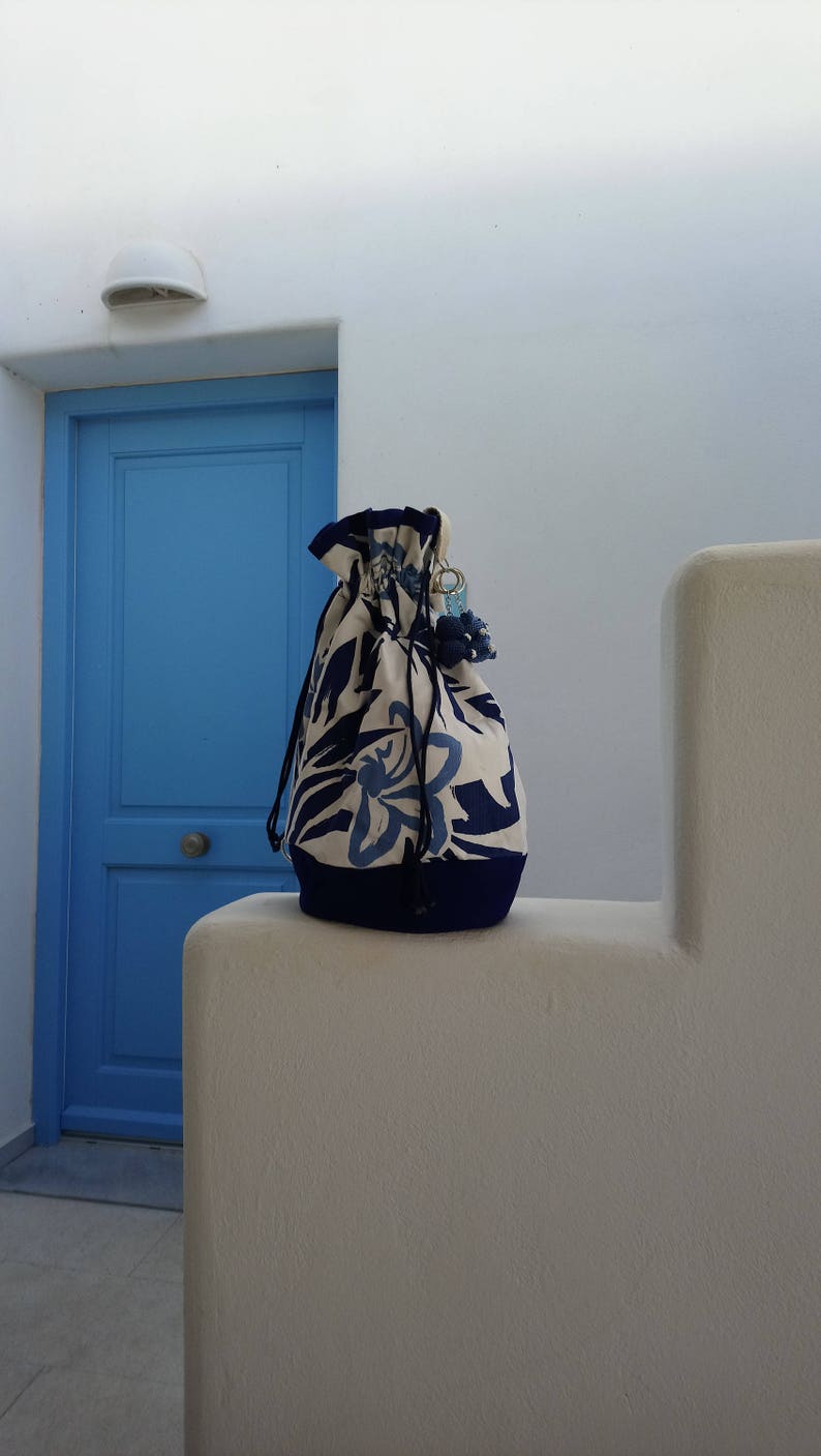 May include: A white drawstring bag with a blue floral pattern and a blue trim. The bag is sitting on a white surface in front of a blue door.
