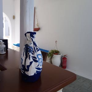 May include: A white and blue floral drawstring bag with a blue and white charm hanging from the top. The bag is sitting on a brown wooden table.