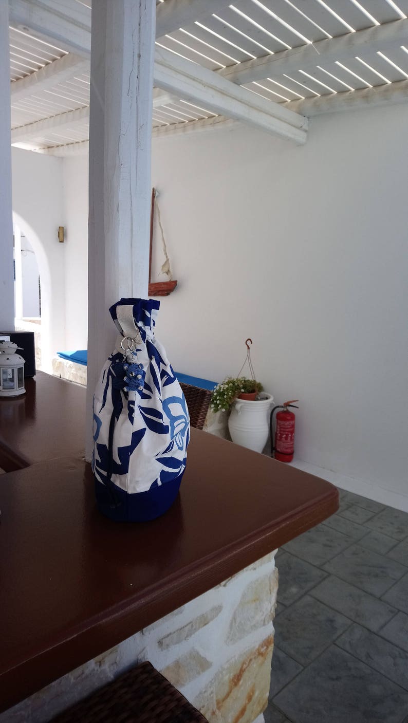 May include: A white and blue floral drawstring bag with a blue and white charm hanging from the top. The bag is sitting on a brown wooden table.