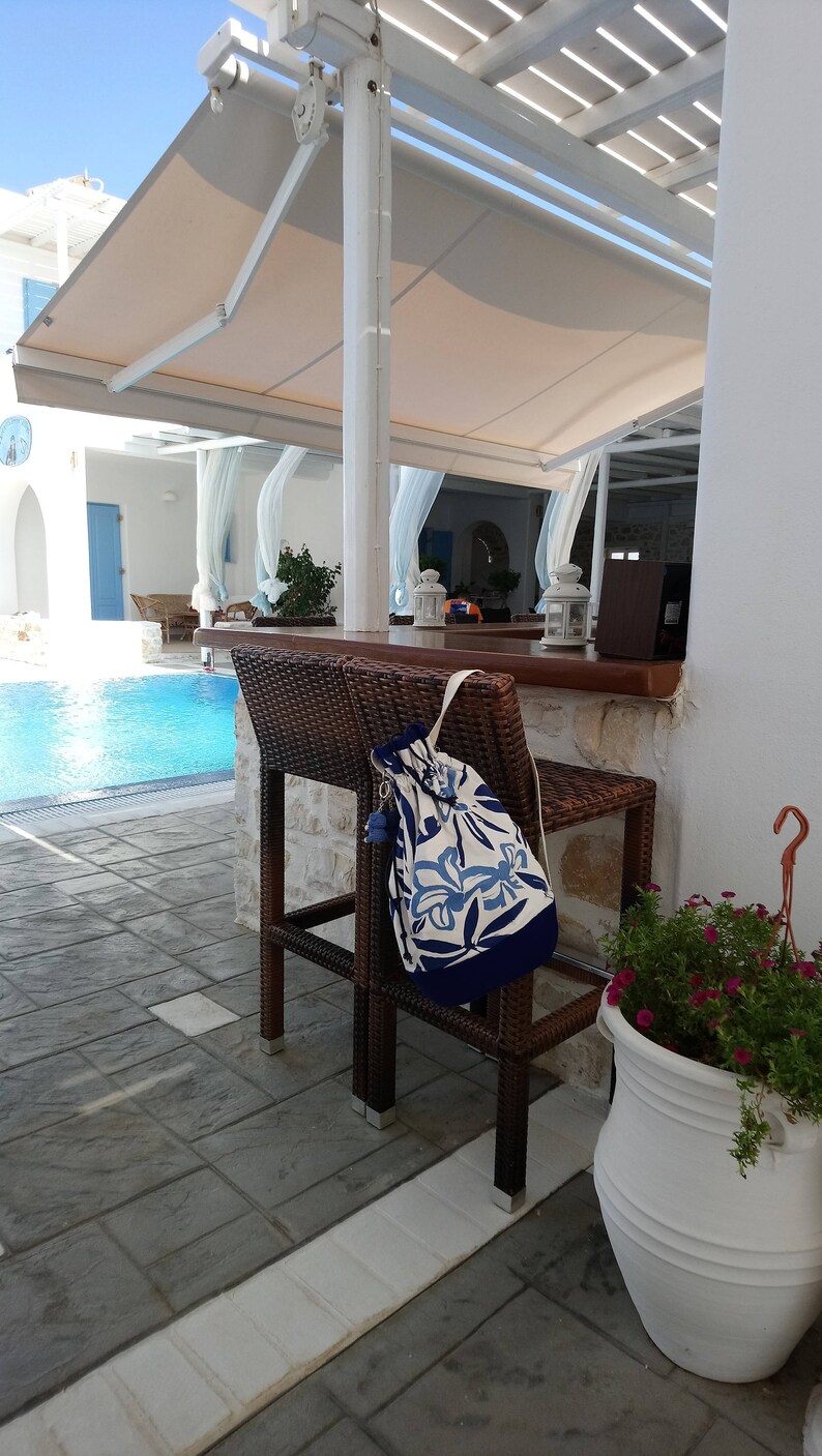 May include: A blue and white patterned drawstring bag hanging on a brown wicker chair by a swimming pool. The chair is on a patio with a white tiled floor.