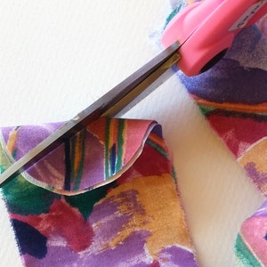 May include: A pair of pink fabric scissors cutting a piece of colourful fabric with a curved edge. The fabric has a floral pattern in shades of pink, purple, green and orange.