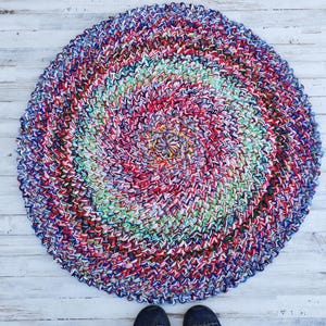 May include: A round, handmade rug with a spiral design. The rug features a vibrant mix of colors, including red, blue, green, and purple. The rug is made of a thick, textured material and is placed on a light-colored wooden floor.