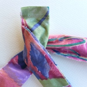 May include: A close-up of a fabric strip with a colourful abstract print in shades of pink, green, blue, and purple. The fabric is folded and tied in a knot, showing the raw edges of the fabric.