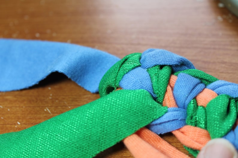 May include: A close-up of a braided fabric toy made from strips of blue, green, and orange fabric. The fabric is woven together to create a knot.