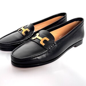 May include: A pair of black leather loafers with gold-colored metal accents. The shoes have a classic design with a slip-on style and a decorative buckle on the vamp. The loafers are set against a white background.