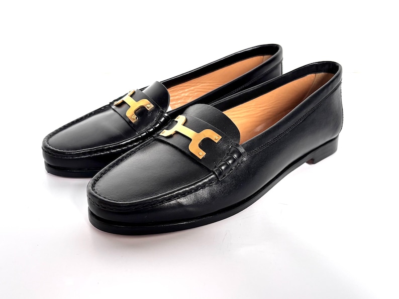 May include: A pair of black leather loafers with gold-colored metal accents. The shoes have a classic design with a slip-on style and a decorative buckle on the vamp. The loafers are set against a white background.