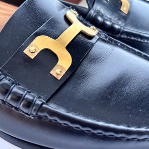 May include: Close-up of black leather loafers with a gold-colored metal accent. The shoes have a classic design with a stitched sole and a decorative gold buckle.