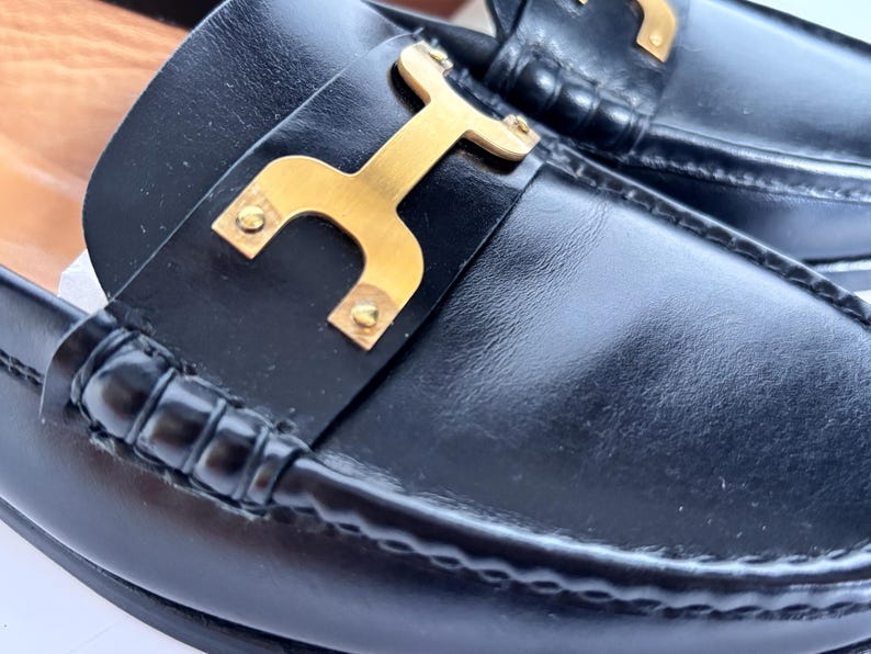 May include: Close-up of black leather loafers with a gold-colored metal accent. The shoes have a classic design with a stitched sole and a decorative gold buckle.