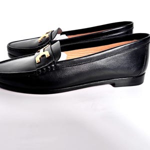 May include: A pair of black leather loafers with a gold-colored accent on the vamp. The shoes have a classic design with a rounded toe and a low heel. The loafers are displayed on a white background.
