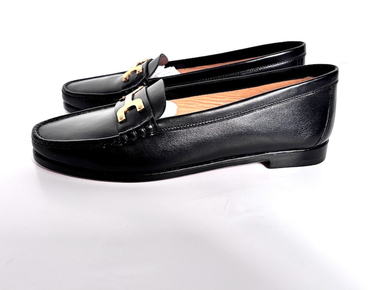 May include: A pair of black leather loafers with a gold-colored accent on the vamp. The shoes have a classic design with a rounded toe and a low heel. The loafers are displayed on a white background.