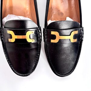 May include: A pair of black leather loafers with gold-colored hardware. The shoes have a classic design with a decorative strap across the top. The loafers are new and have white paper inside.