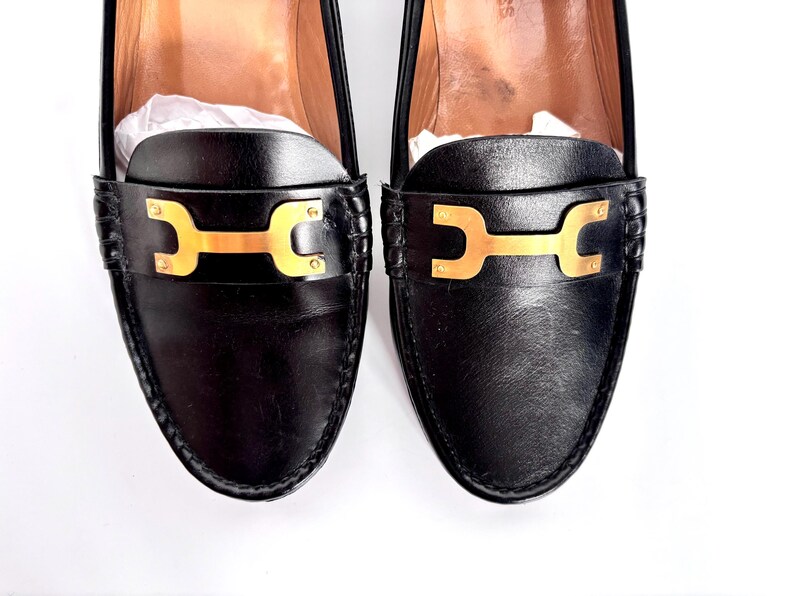 May include: A pair of black leather loafers with gold-colored hardware. The shoes have a classic design with a decorative strap across the top. The loafers are new and have white paper inside.