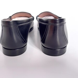 May include: A pair of black leather loafers, viewed from the back. The shoes have a classic design with a rounded toe and a low heel. The leather appears smooth and polished, with visible stitching details. The interior of the shoes is a light tan color.
