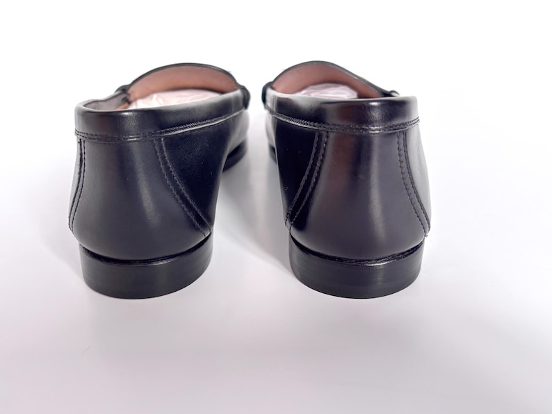 May include: A pair of black leather loafers, viewed from the back. The shoes have a classic design with a rounded toe and a low heel. The leather appears smooth and polished, with visible stitching details. The interior of the shoes is a light tan color.