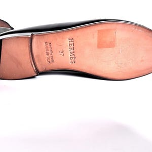 May include: Black leather shoe with a light brown sole. The sole is stamped with the brand name "HERM&Egrave;S", the size "37", and "Semelle cuir Made in Italy". The shoe is on a white background.