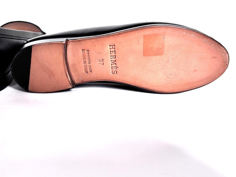 May include: Black leather shoe with a light brown sole. The sole is stamped with the brand name "HERM&Egrave;S", the size "37", and "Semelle cuir Made in Italy". The shoe is on a white background.