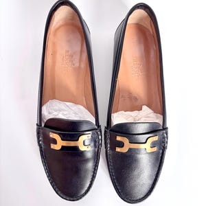 May include: A pair of black leather loafers with gold-colored hardware. The shoes have a classic design with a rounded toe and a low heel. The interior of the shoes is a light tan color, and the brand name is visible.