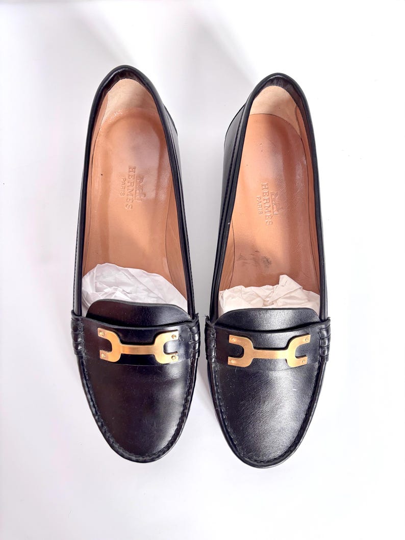 May include: A pair of black leather loafers with gold-colored hardware. The shoes have a classic design with a rounded toe and a low heel. The interior of the shoes is a light tan color, and the brand name is visible.