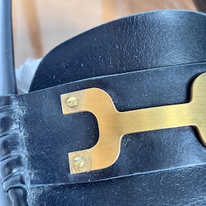 May include: Close-up of a black leather shoe with a gold-colored metal accent. The metal piece is shaped like a stylized letter "H" and is attached to the shoe with small gold rivets. The leather appears to be slightly worn.