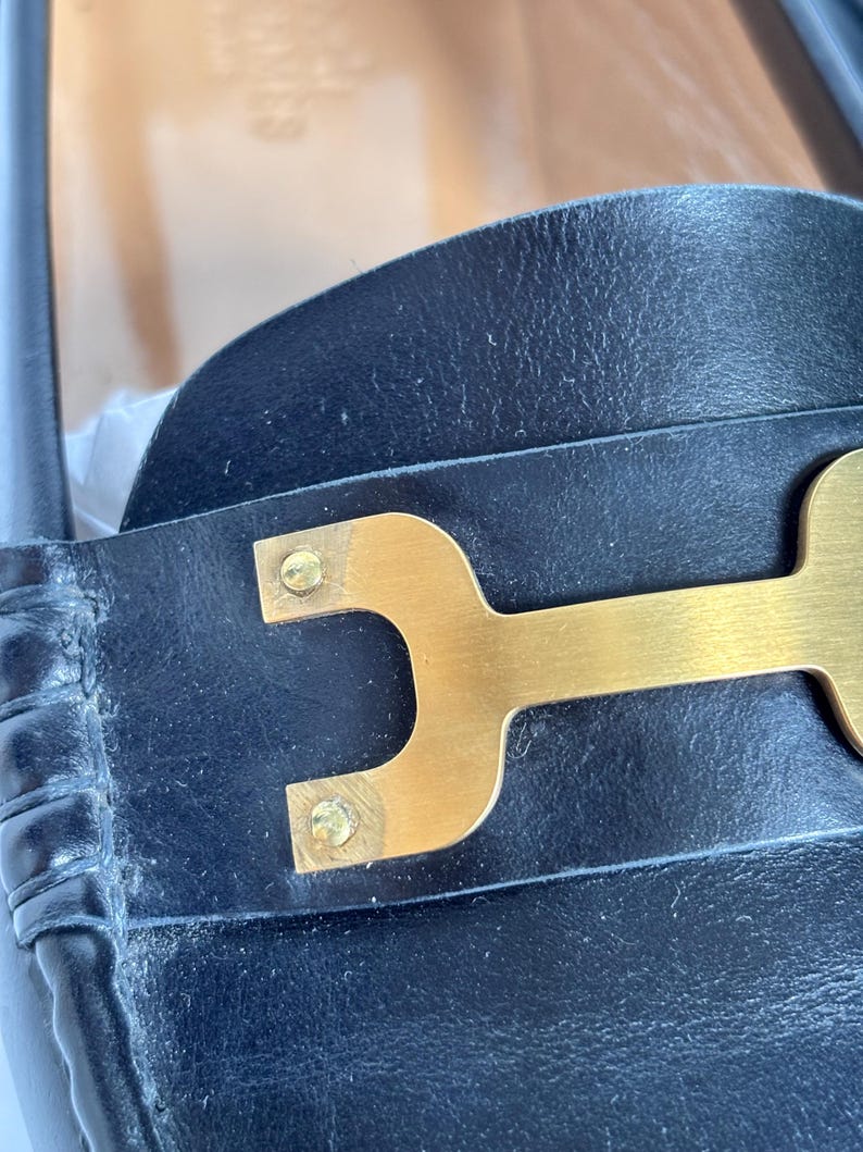 May include: Close-up of a black leather shoe with a gold-colored metal accent. The metal piece is shaped like a stylized letter "H" and is attached to the shoe with small gold rivets. The leather appears to be slightly worn.
