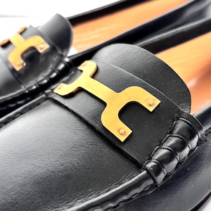 May include: Close-up of a pair of black leather loafers. Each shoe features a gold-colored metal accent in the shape of a stylized letter "H" on the vamp. The shoes have a classic, elegant design, suitable for various occasions.