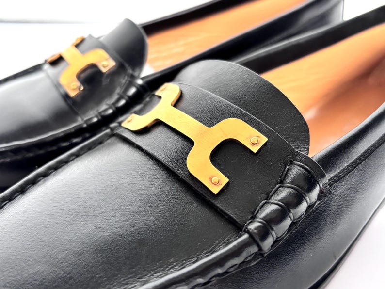 May include: Close-up of a pair of black leather loafers. Each shoe features a gold-colored metal accent in the shape of a stylized letter "H" on the vamp. The shoes have a classic, elegant design, suitable for various occasions.