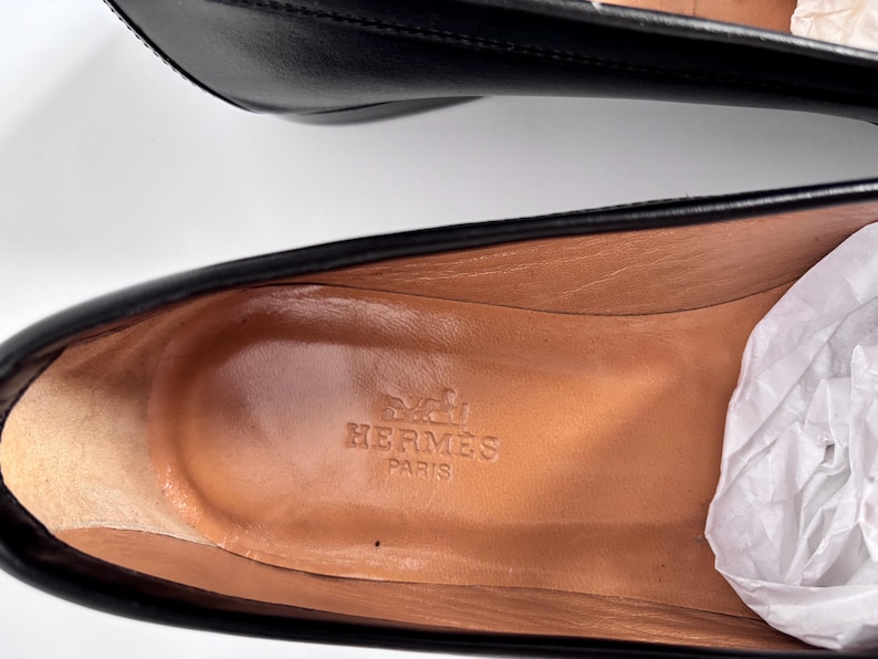 May include: Close-up of a pair of black leather shoes. The interior of one shoe is visible, showcasing a tan insole with the embossed "Herm&egrave;s Paris" logo. White tissue paper is partially visible.