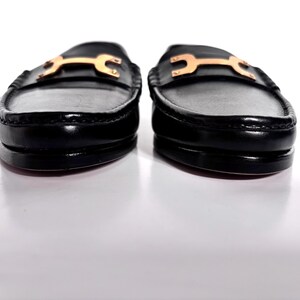 May include: Pair of black leather loafers with gold-colored hardware. The shoes have a classic design with a rounded toe and a low heel. The gold hardware adds a touch of elegance to the footwear.