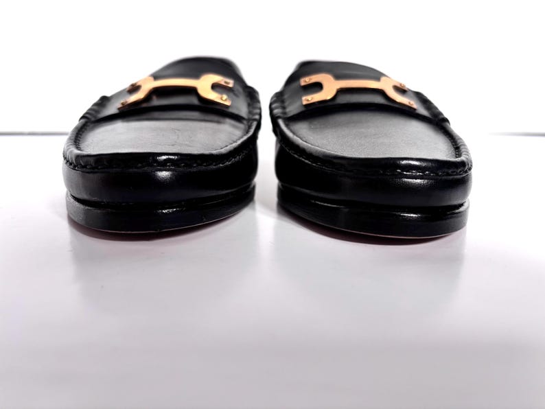 May include: Pair of black leather loafers with gold-colored hardware. The shoes have a classic design with a rounded toe and a low heel. The gold hardware adds a touch of elegance to the footwear.