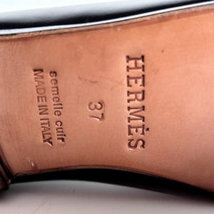 May include: Close-up of a black leather shoe sole with the brand name "HERM&Egrave;S" embossed in brown. The size "37" is visible, along with text indicating "semelle cuir" and "MADE IN ITALY".