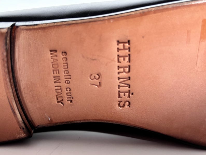 May include: Close-up of a black leather shoe sole with the brand name "HERM&Egrave;S" embossed in brown. The size "37" is visible, along with text indicating "semelle cuir" and "MADE IN ITALY".