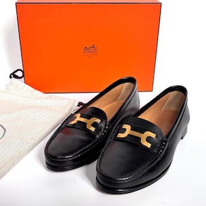 May include: A pair of black leather loafers with gold-tone hardware. The shoes are displayed with a matching orange box and a dust bag. The loafers have a classic design, suitable for various occasions.