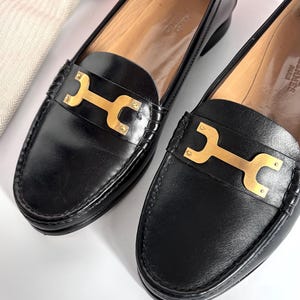 May include: A pair of black leather loafers with gold-tone hardware. The shoes feature a classic design with a rounded toe and a decorative gold bar across the vamp. The interior is a light tan color. A small, off-white drawstring bag is in the background.