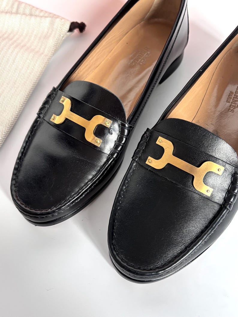 May include: A pair of black leather loafers with gold-tone hardware. The shoes feature a classic design with a rounded toe and a decorative gold bar across the vamp. The interior is a light tan color. A small, off-white drawstring bag is in the background.