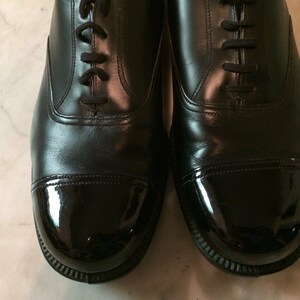 May include: A pair of black leather shoes with a cap toe. The cap toe is a shiny black patent leather.