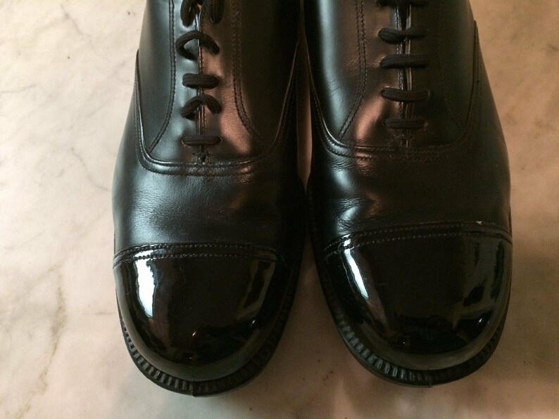 May include: A pair of black leather shoes with a cap toe. The cap toe is a shiny black patent leather.