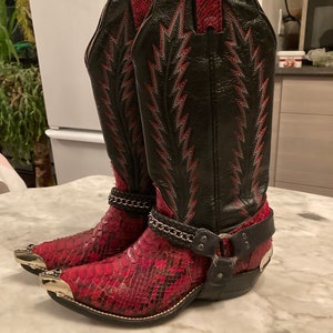 Peut inclure: Une paire de bottes de cow-boy rouges et noires avec un motif en peau de serpent et des accents argentés. Les bottes ont une pointe pointue et un talon haut.