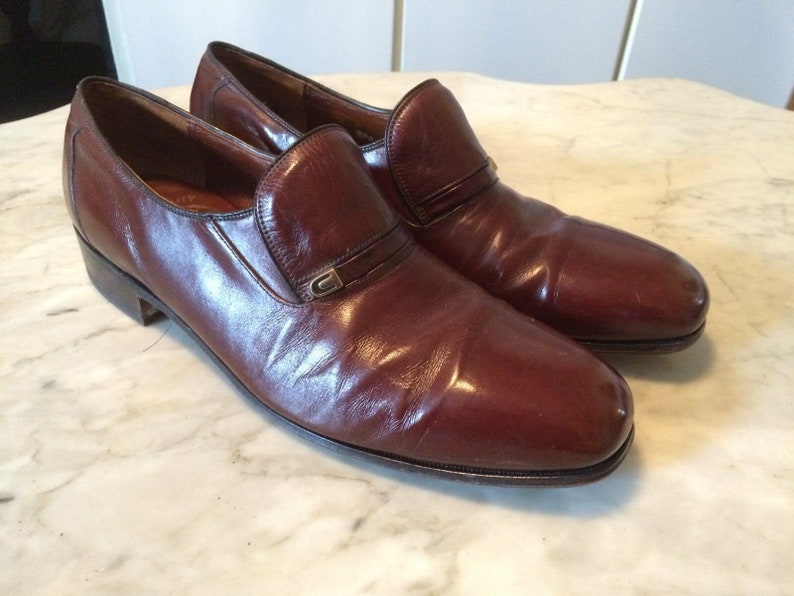 May include: A pair of brown leather slip-on shoes with a buckle detail on the front.